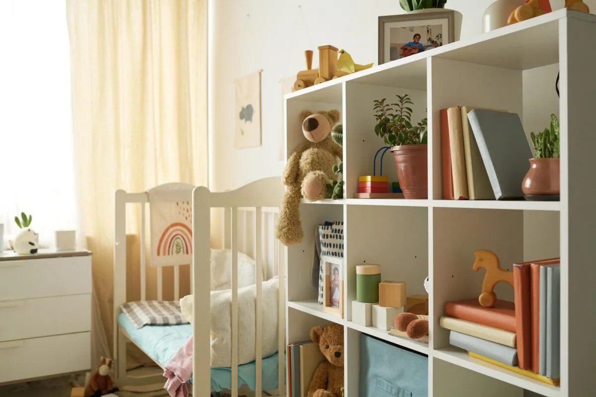 Estante de nichos brancos com livros, brinquedos de madeira e ursos de pelúcia ao lado de um berço em quarto de bebê