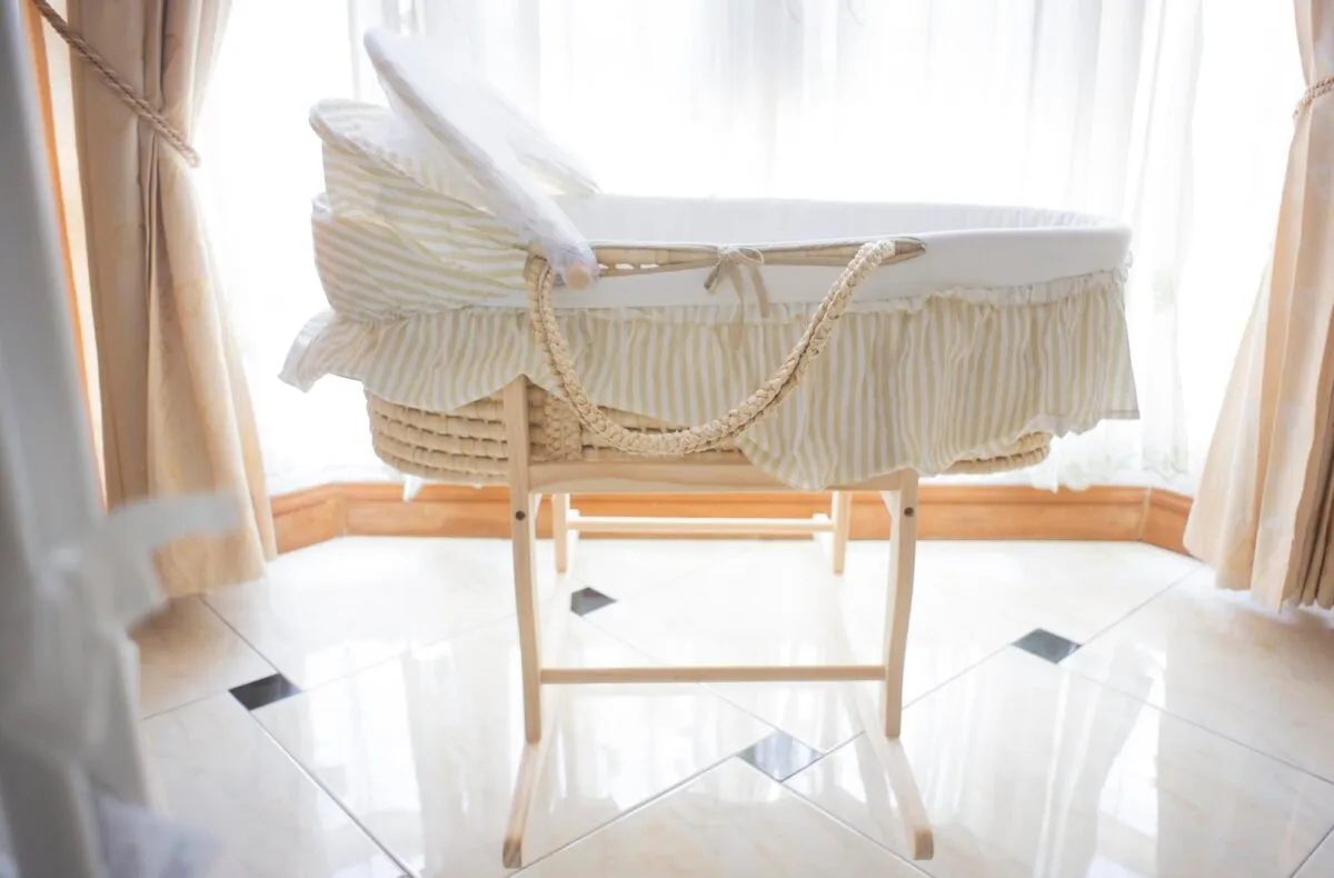 Moisés portátil de palha com saia listrada sobre suporte de madeira dobrável, em quarto claro com cortinas e janela ao fundo