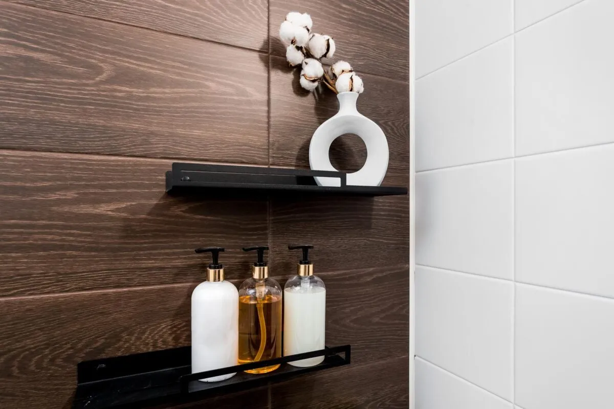 Prateleiras pretas em parede de azulejo amadeirado com frascos de shampoo e vaso branco com flor de algodao