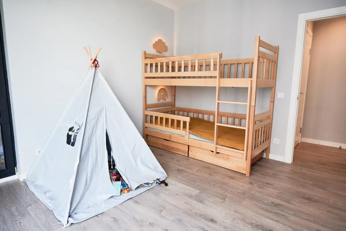 Quarto infantil com beliche de madeira com grade de proteção, cabana de brincar cinza e piso laminado, iluminado por luz natural
