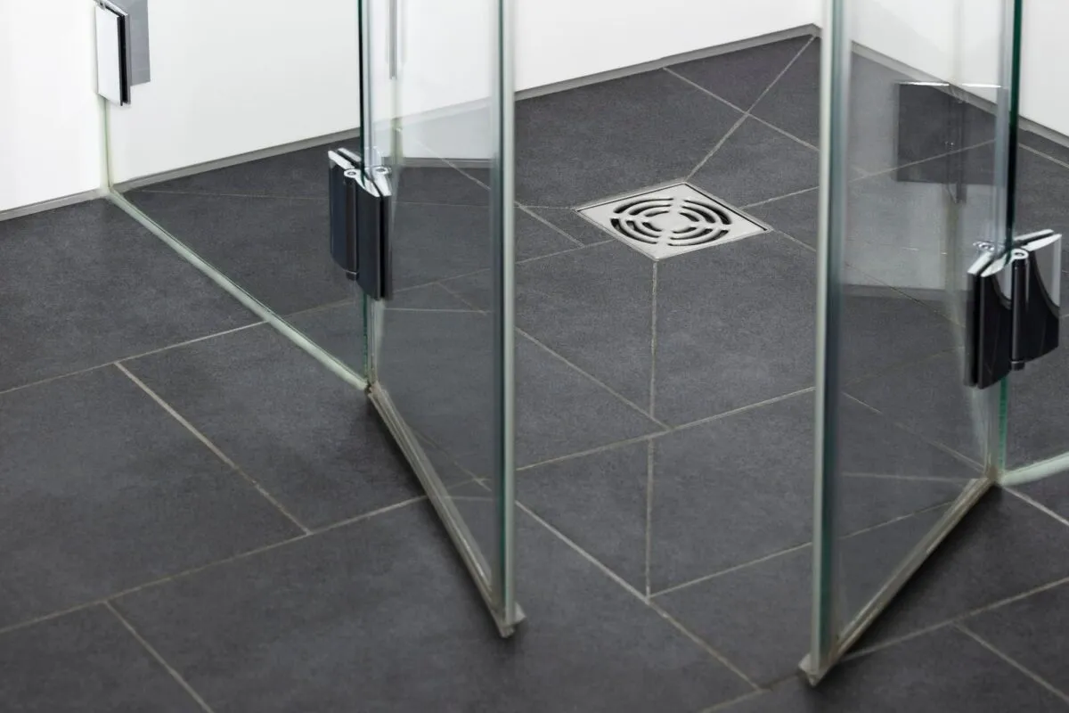 Ralo de chão quadrado em aço inox no piso de porcelanato cinza de um box de banheiro com divisórias de vidro