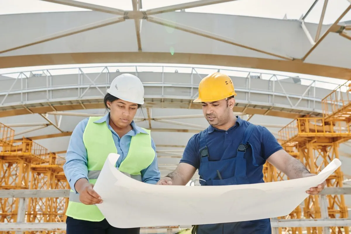 Engenheira e profissional de obra conferem a planta impressa no canteiro, com estrutura metálica e guindastes ao fundo